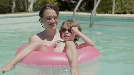 cheerful little boy floating on inflatable ring in swimming pool with the help of his brother and posing for camera