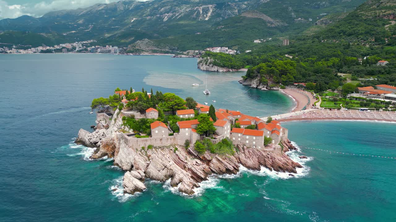 Sveti Stefan, Montenegro. Aerial drone view of famous and luxury travel destination. Static footage. Tourist boat passing. At right Sveti Stefan Beach.