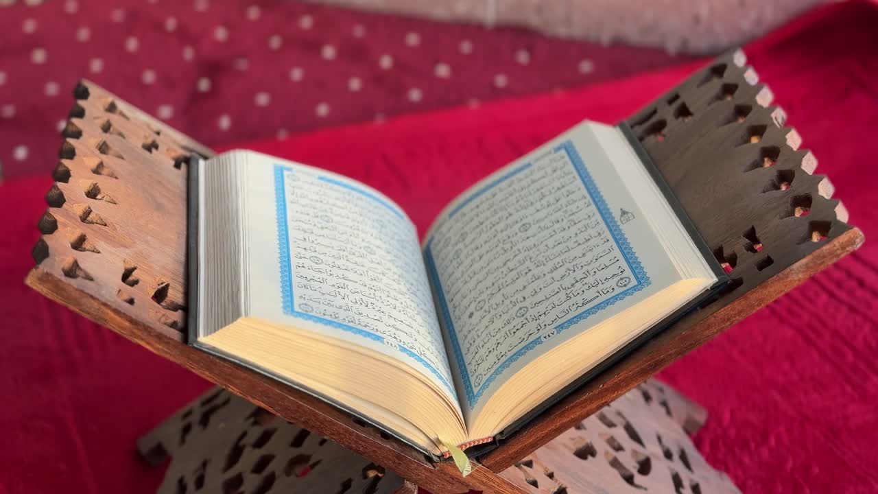 4K close-up of the Holy Quran open on a traditional wooden rehal over a red prayer rug. Ideal for spiritual, cultural, or religious-themed video content and documentaries.
