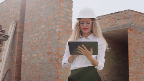 arquitecta femenina en ropa de negocios y un casco blanco se encuentra en una casa de nueva construcción con paredes no tratadas y trabaja en una tableta. tecnologías modernas en las profesiones más antiguas