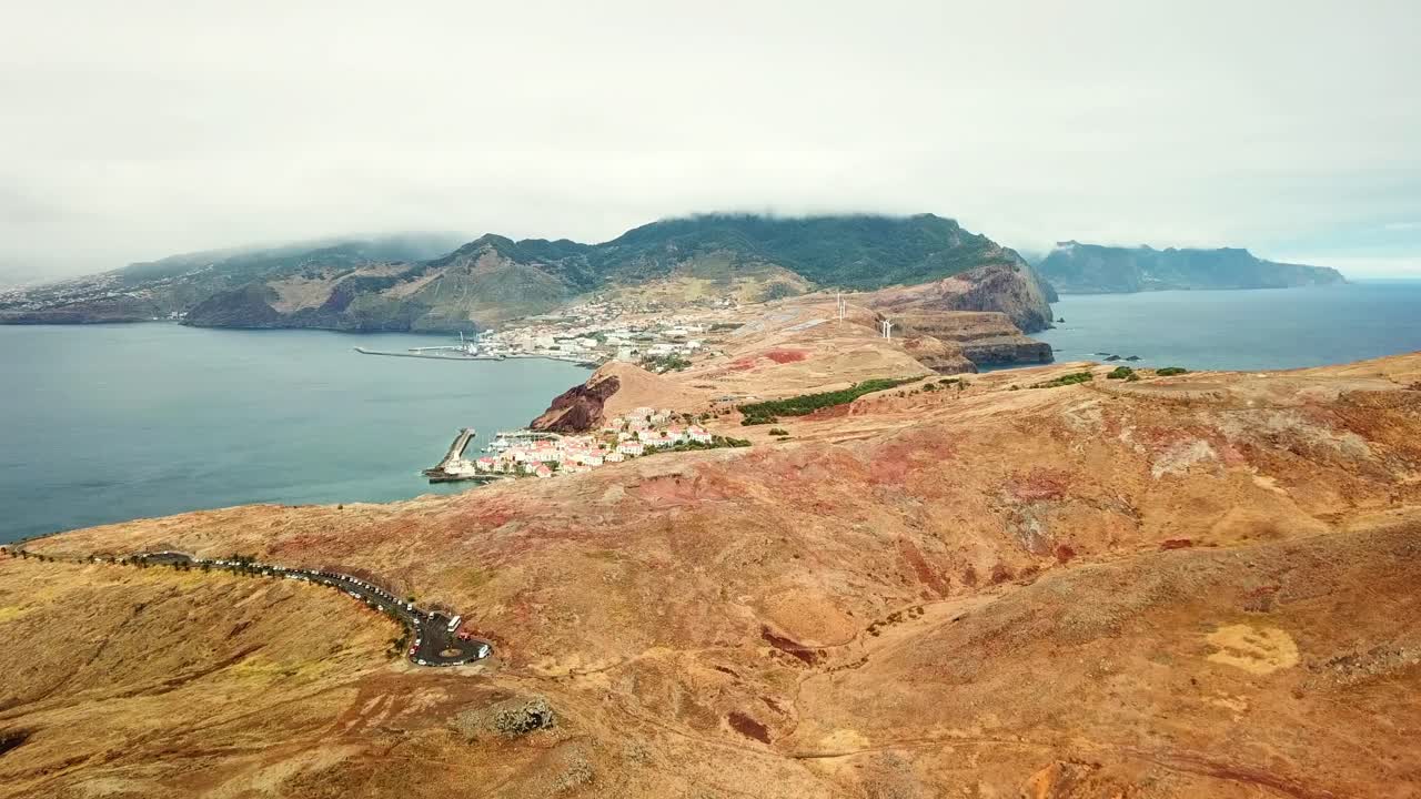 Aerial view of Ponta de São Lourenço in Madeira, highlighting rugged cliffs, tranquil ocean waters, and barren landscapes. Features a coastal village and stunning natural terrain under a cloudy sky
