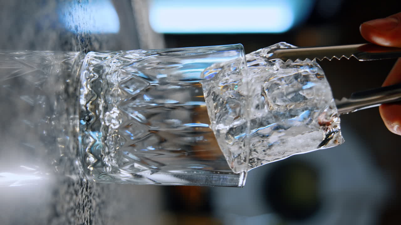 Large ice cube is placed into a glass using the tweezers. Preparing the drink in the bar. Close up. Vertical screen. Blurred backdrop.