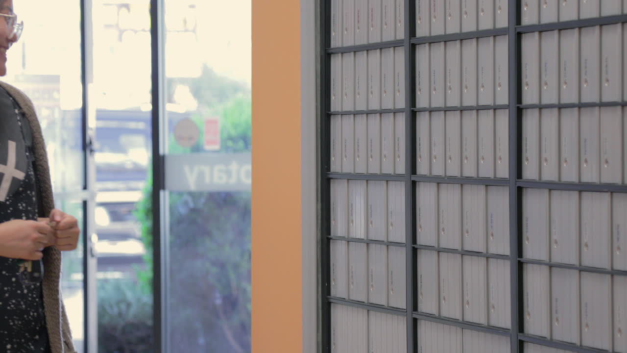 안경을 쓴 젊은 여성이 우체국에 들어가 우편물을 확인하기 위해 우편함으로 걸어갑니다.