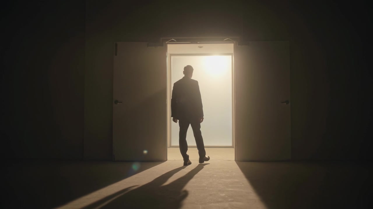 Uomo in abito che cammina, fermandosi davanti a luminose porte a doppio battente in una stanza in penombra, controluce che lo delinea, lunga ombra