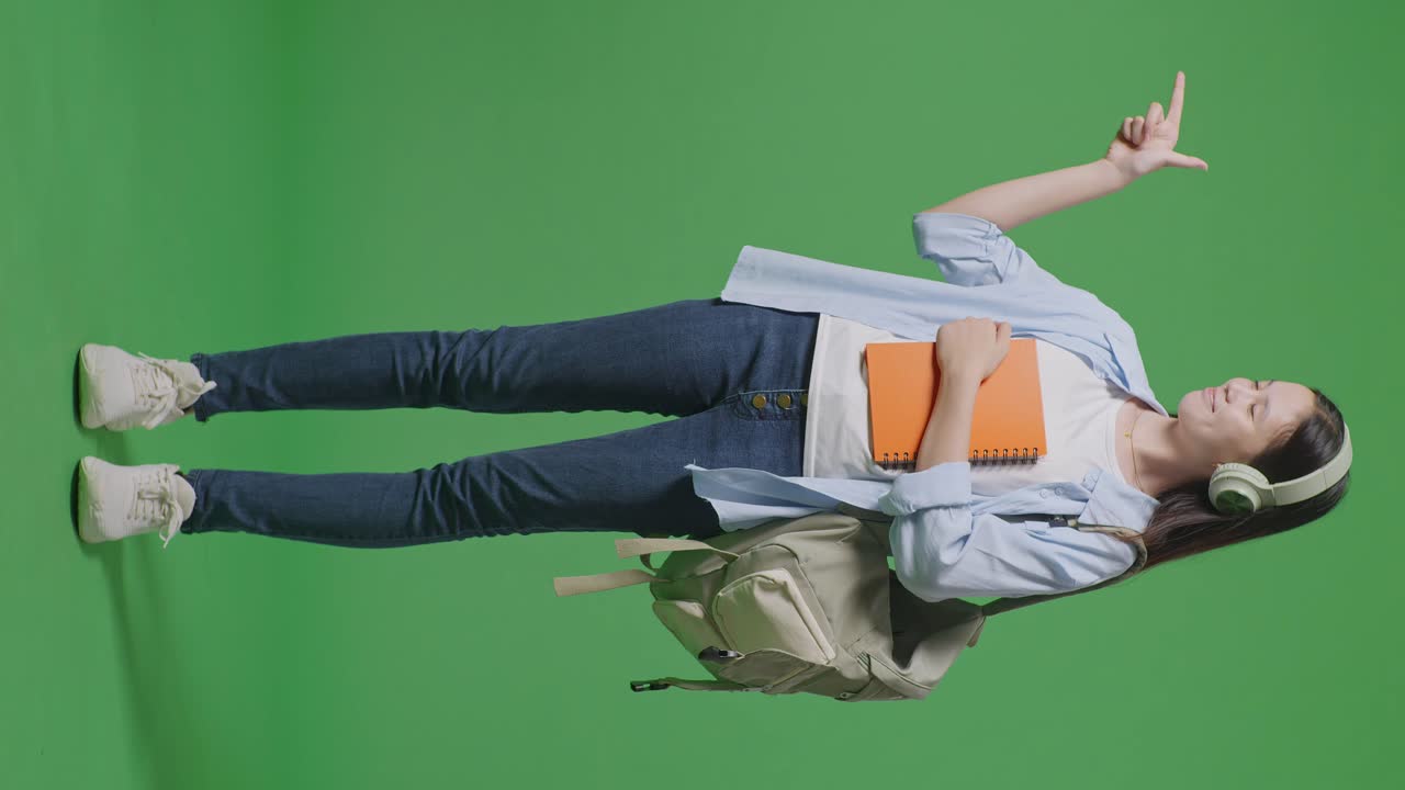cuerpo lleno de estudiante adolescente asiática con una mochila y algunos libros sonriendo y señalando a un lado mientras estaba de pie en el estudio de fondo de la pantalla verde