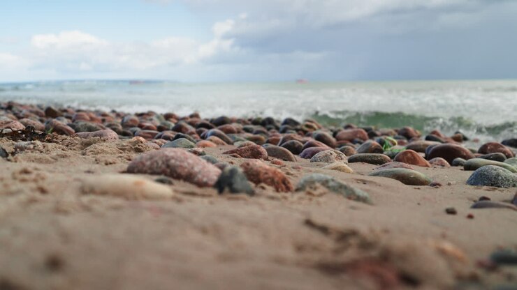 Baltic sea water calm waves crashing against shore covered in pebble stones making splashes, sunny summer day in Rugen Island - slow motion low angle