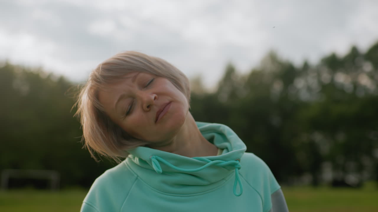 Sport woman in mint tracksuit performing neck rotation exercise in open green field under cloudy sky during morning workout session focusing on relaxation stretching flexibility and fitness routine