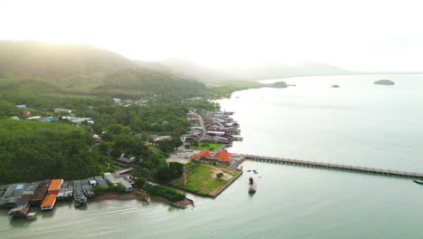 Aerial-backwards-shot-of-Koh-Lanta-coastline-with-jetty-and-green-mountain-landscape-at-sunrise,-Thailand