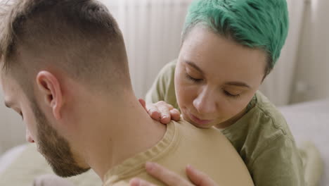 young woman touching her boyfriend's head and then resting on his shoulder while sitting together on the bed at home