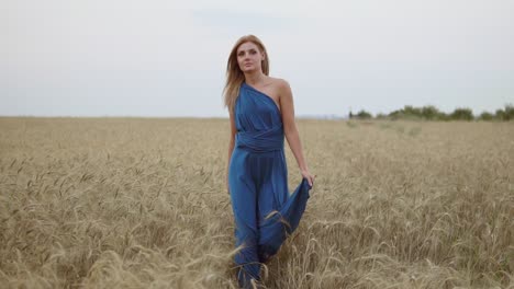 Hermosa-Joven-Feliz-Con-Un-Largo-Vestido-Azul-Caminando-Por-Un-Campo-De-Trigo-Dorado-Mirando-A-La-Cámara.-Concepto-De-Libertad