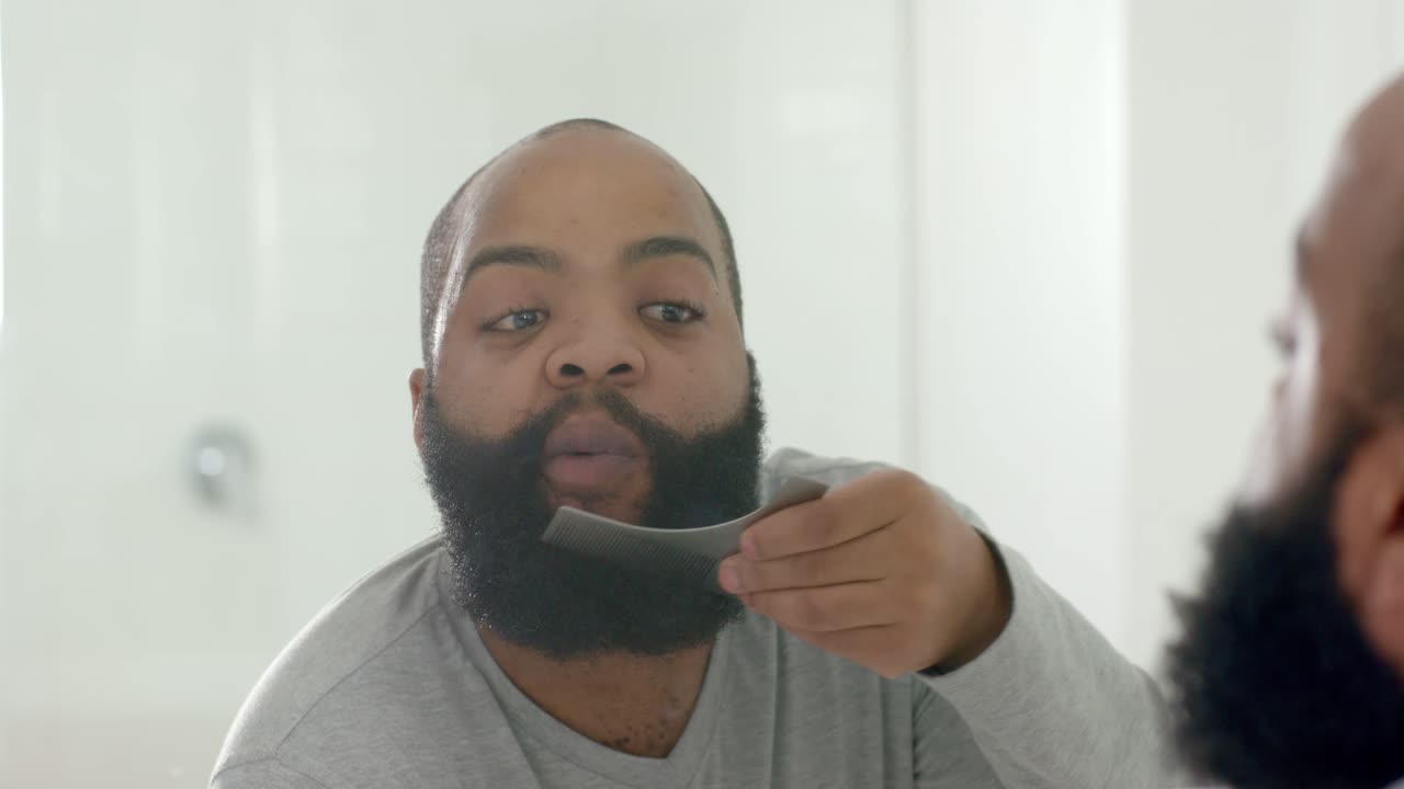 Man checking bathroom mirror, holding wide comb, combing beard and noting scissors for symmetry