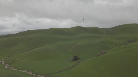 drone pulls out overlooking a large grassy hill with windmills on overcast day