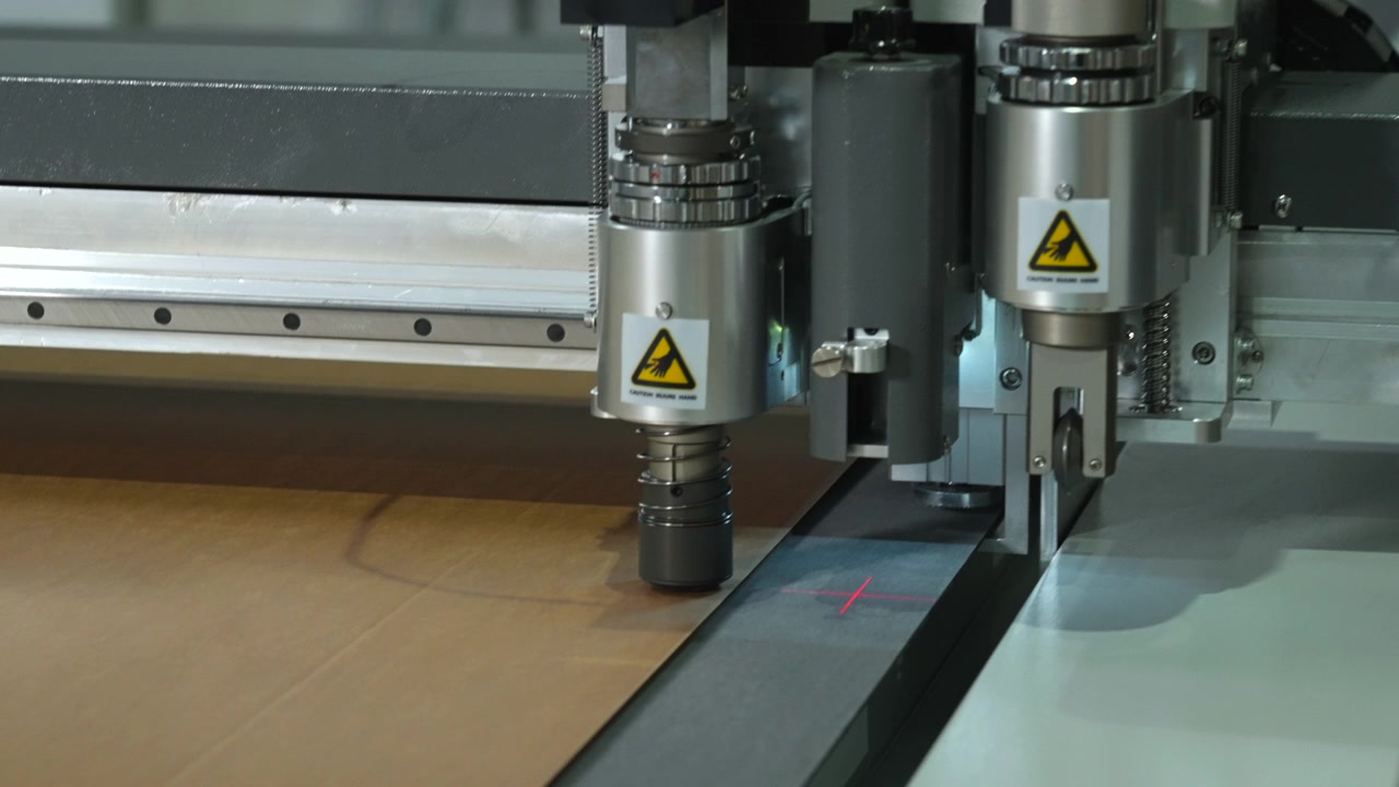 Cabeçote de máquina de corte industrial desliza sobre material de folha marrom dentro de oficina, mostrando trilhos metálicos, peças-guia, adesivos de aviso e marcas de alinhamento brilhantes em superfície lisa com luz uniforme