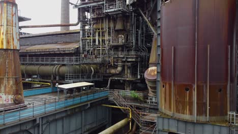 overview-of-rusty-warehouse-factory-with-ladder-metal-constructions-tubes-and-pipes-connected-fire-exit-steps-leading-to-the-top-of-the-building-power-plant-with-smokestack-at-the-background-Germany