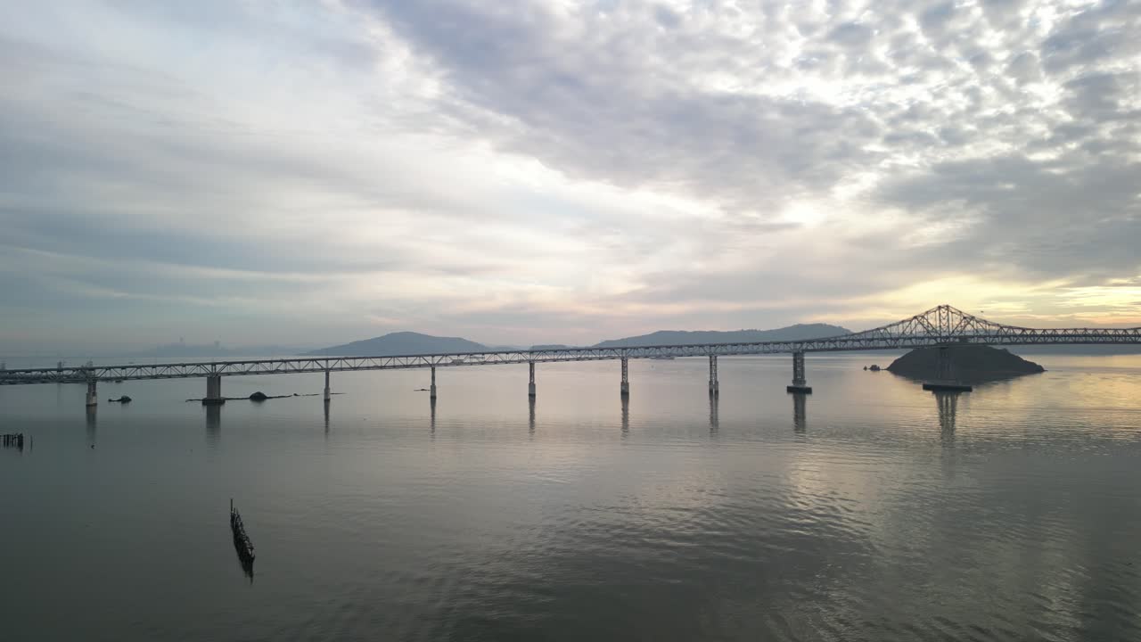 Aerial drone footage at sunset captures the Richmond–San Rafael Bridge stretching across the bay, with moving traffic and shimmering waters below.