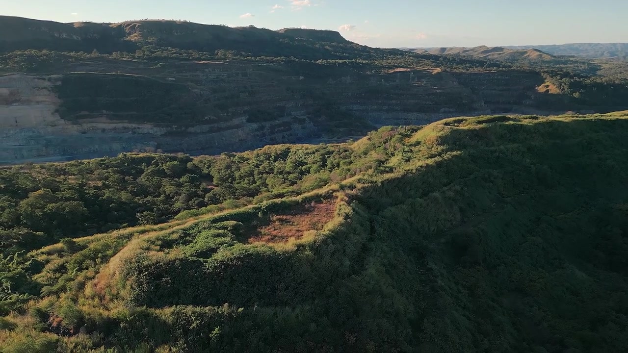 Movimento laterale di drone dall'alto che si sposta sopra una cresta illuminata dal sole coperta da vegetazione nativa del Cerrado, rivelando sullo sfondo una vasta cava di calcare a terrazze a Fercal, Brasilia, Brasile.