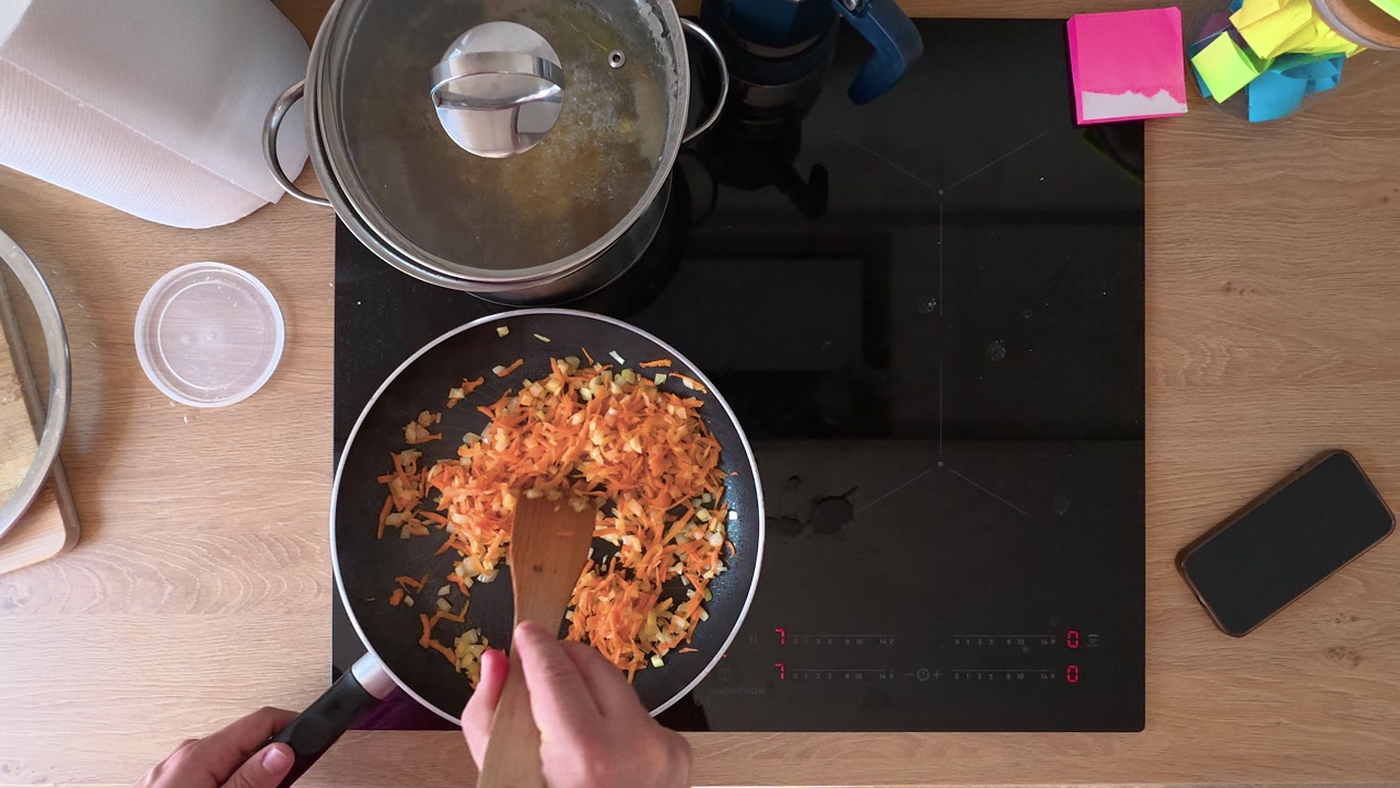A close-up shot showing hands stirring grated vegetables in a skillet on a modern stovetop. The warm kitchen lighting creates an inviting atmosphere, emphasizing cooking ingredients.