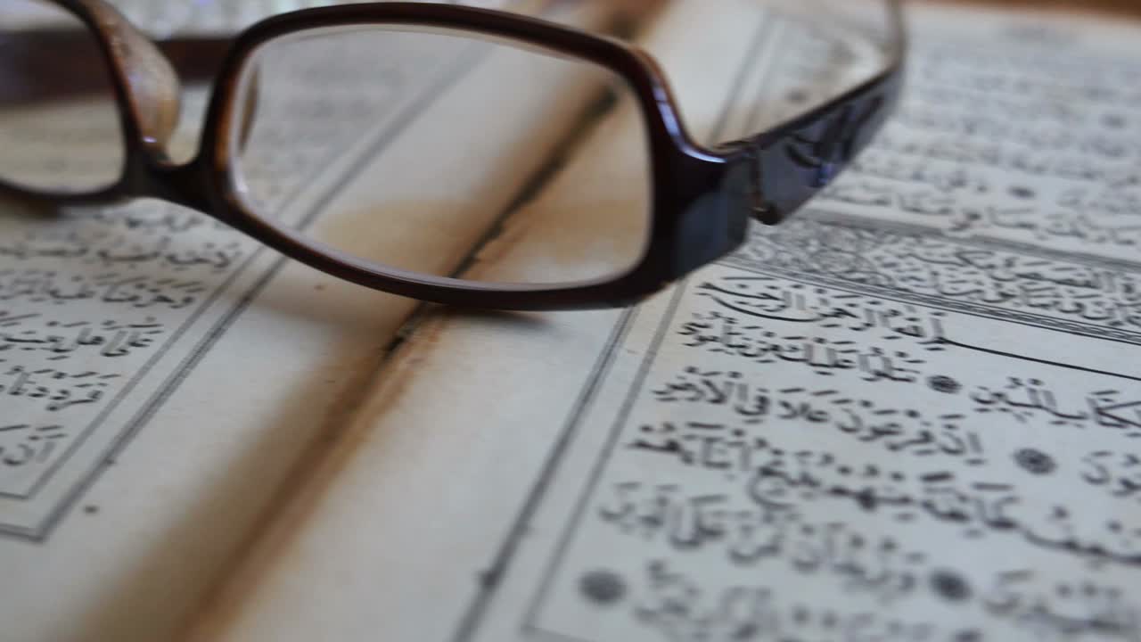 Footage of a Quran lying on a table with reading glasses on it and a tasbeeh in the back, the shot is moving from the left to the right