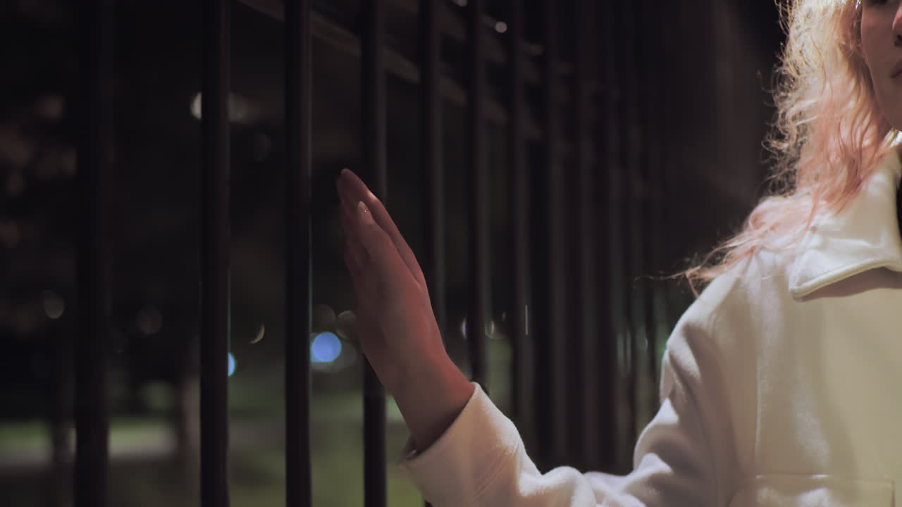 Partial view of woman wearing white coat gently gliding hand over iron fence at night, illuminated by soft ambient lighting with colorful bokeh effect in background