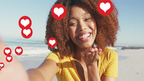 animation of red heart love digital icons over smiling woman blowing kisses on beach