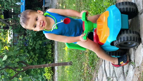 caucasian child in blue t-shirt and pants plays outdoors. baby boy sits on toy car looking aside and talking. vertical screen.