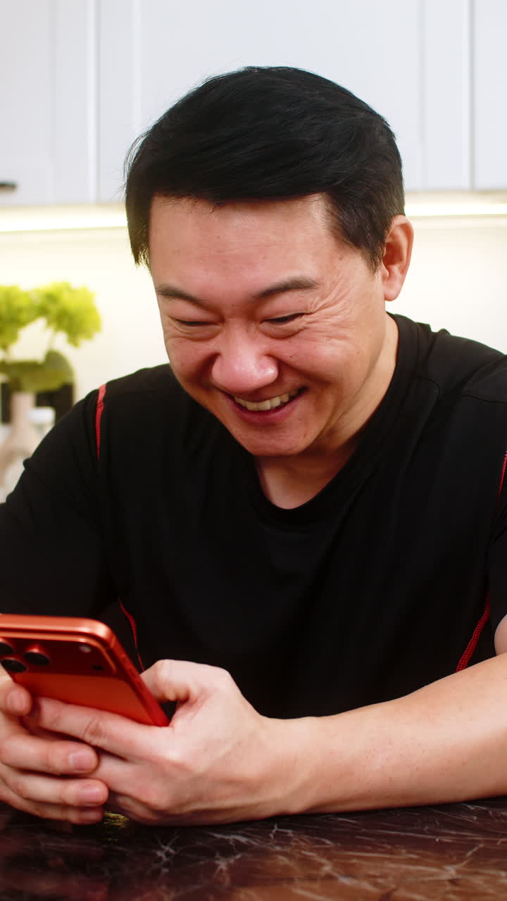 Happy asian man celebrates online game win at home kitchen with orange smartphone feeling excited
