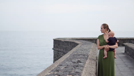 alongside the ocean on a historic european square, a young mother walks with her baby son, watching the waves and exchanging smiles of happiness