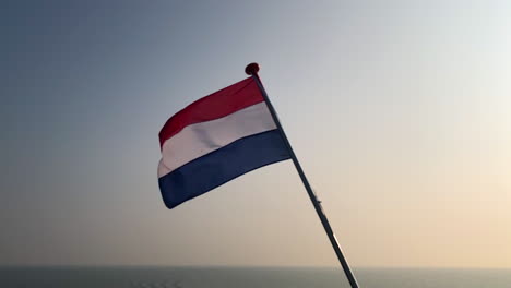 cámara lenta de la bandera holandesa de los países bajos en el viento al atardecer sobre un fondo de agua en movimiento desde el mar del norte