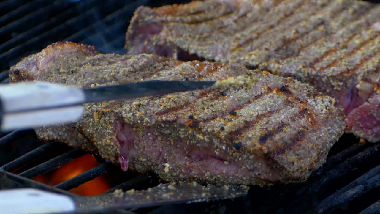un par de pinzas para carne empujan un jugoso filete de costilla casi cocinado en una parrilla y lo deslizan un poco a cámara lenta