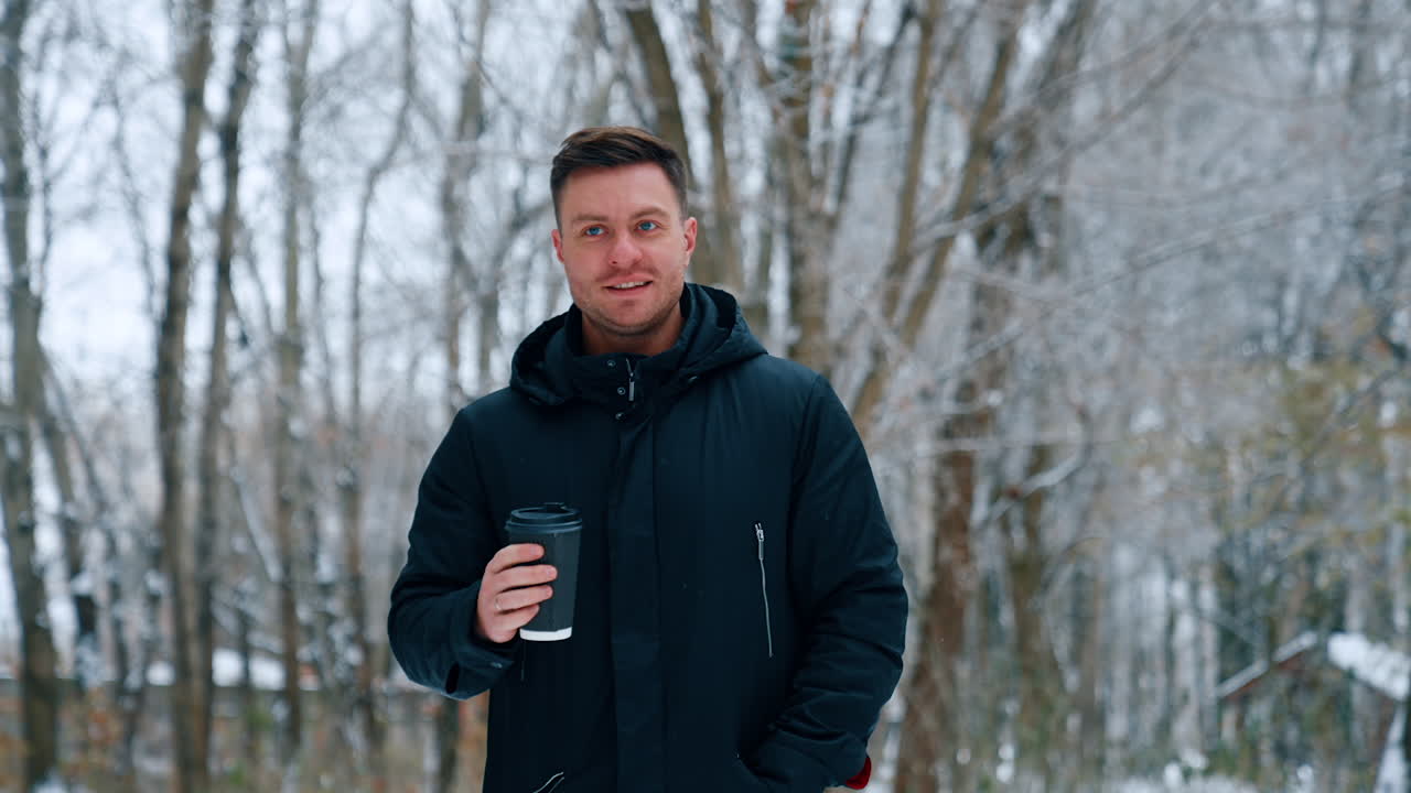 Man wearing warm black winter jacket walks outdoors carrying a paper cup. Man drinks coffee walking in winter park.