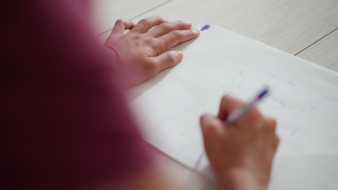 Primer plano de un niño dibujando en un papel colocado en el suelo con una cuerda roja en la muñeca, mostrando un dibujo creativo sencillo con trazos de lápiz y palabras, con movimientos de mano muy precisos.
