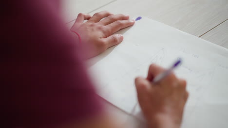 close up of kid wearing red rope around wrist sketching on paper placed on floor showing simple creative drawing with pencil strokes and words while focused hand movements