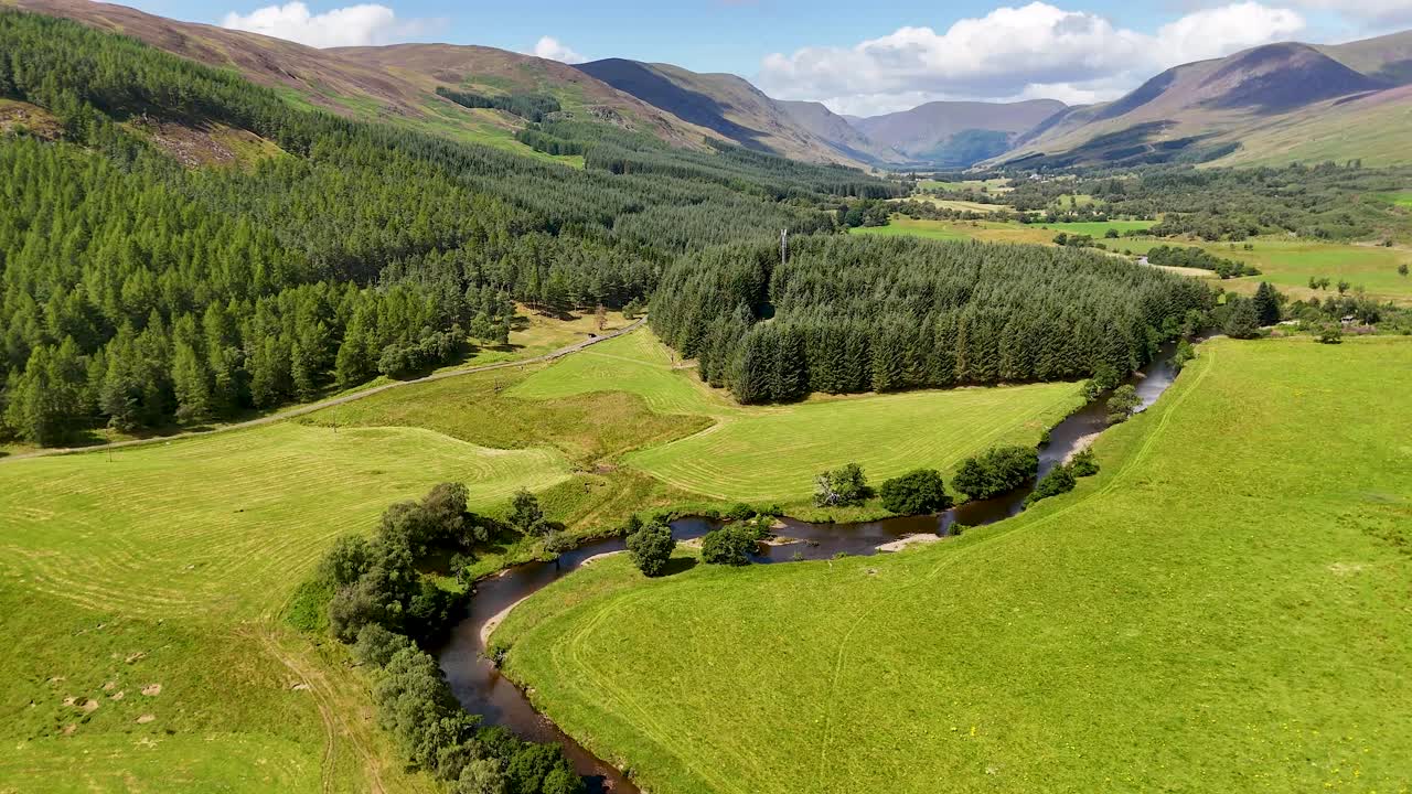 Drone camera glides above a winding river, dense pine forest, and green valley in Glen Clova, Scotland, under bright daylight with clear skies