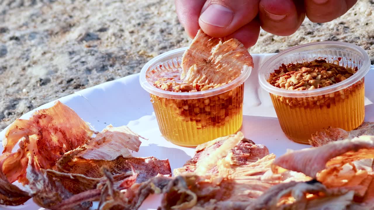 Close-up of a hand dipping dried squid into spicy sauce cups on a paper plate.