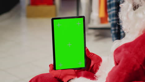 Mitarbeiter-In-Einem-Festlich-Geschmückten-Bekleidungsgeschäft,-Das-Als-Weihnachtsmann-Verkleidet-Ist,-Hält-Ein-Chroma-Key-Tablet-In-Der-Hand,-Richtet-Website-Bekleidungsartikel-Ein-Und-Gibt-Online-Werbeaktionen-Für-Weihnachten-Ein