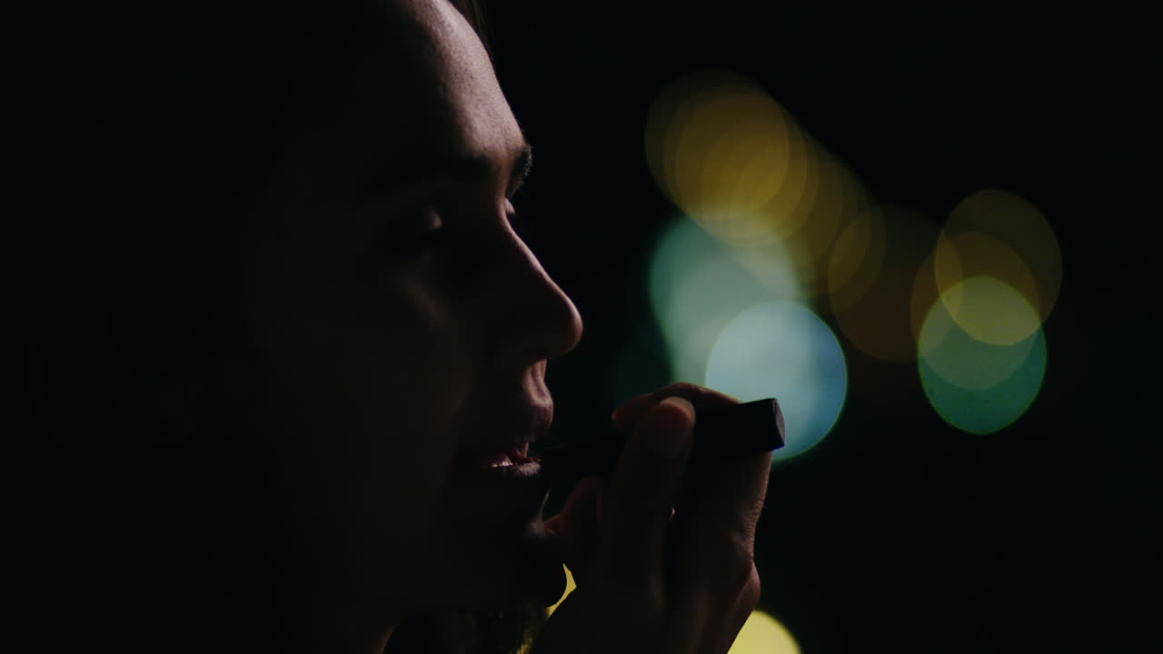 밤에 도시에서 립스틱 메이크업을 하는 아름다운 젊은 여성이 배경에 보케 조명과 함께 저녁 파티를 준비하고 있습니다.