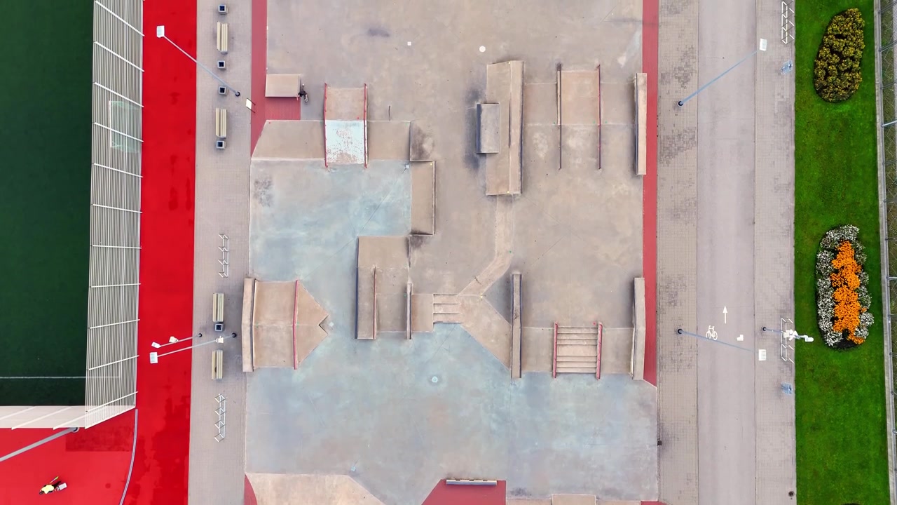 Straight-down drone view of an empty skatepark with concrete ramps, rails and stairs beside a bike lane, red paving and landscaped flower beds.