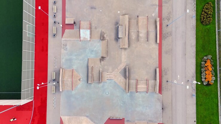Straight-down drone view of an empty skatepark with concrete ramps, rails and stairs beside a bike lane, red paving and landscaped flower beds.