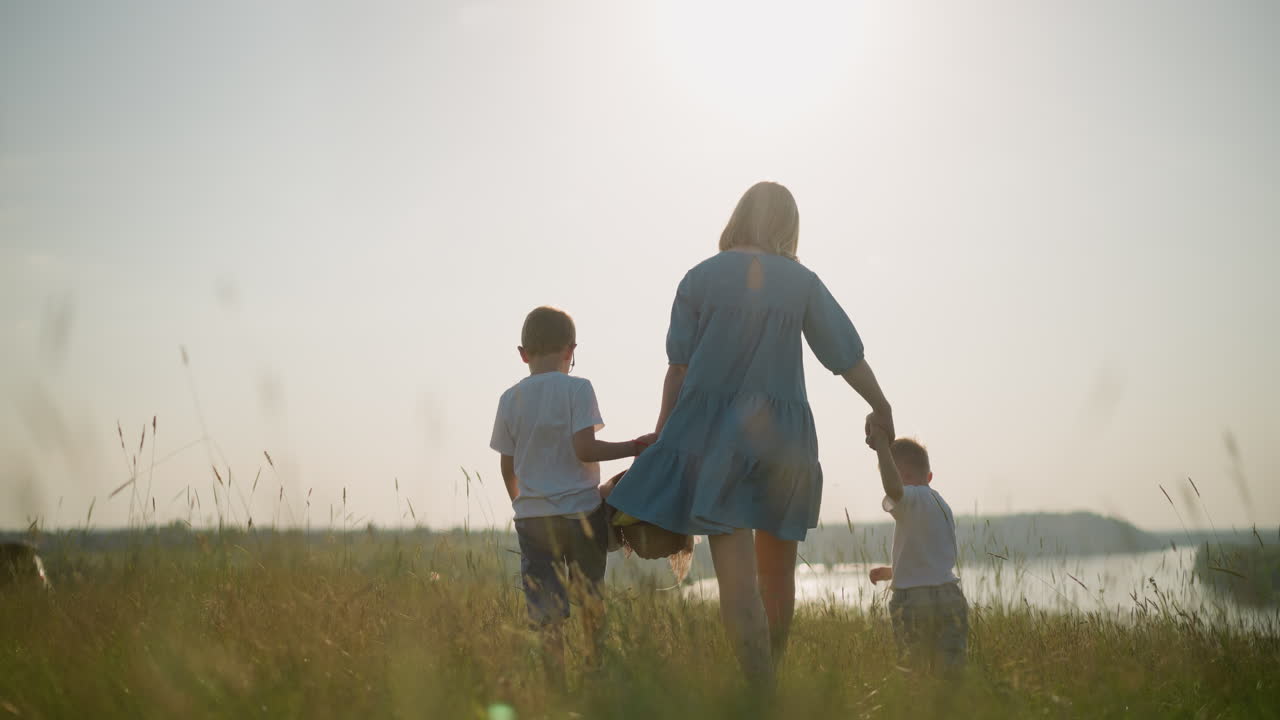 A back shot of a mother in a blue dress carrying a basket in one hand holding her younger son's hand with the other. Her older son walks beside them as they walk through a grassy field towards a lake
