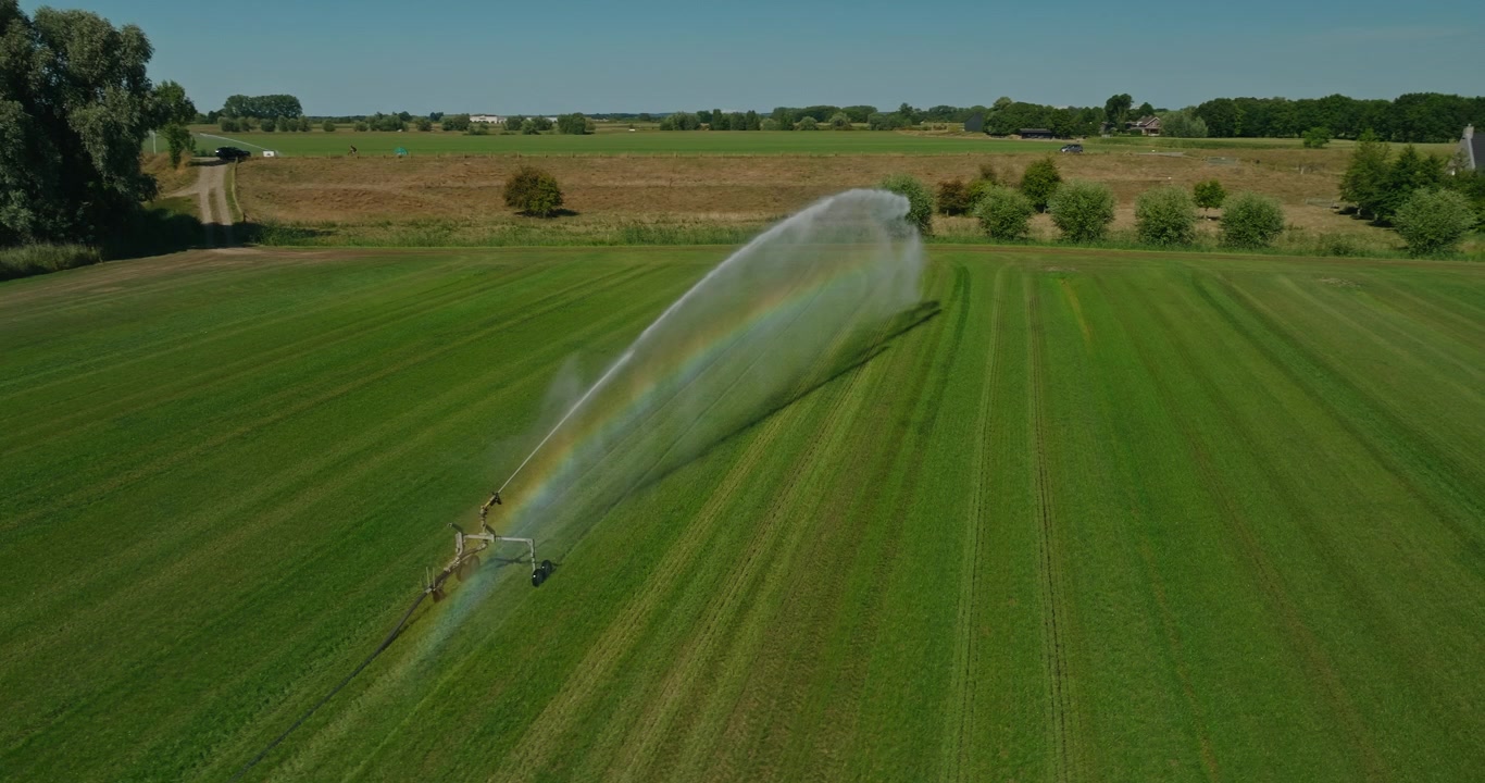Un drone orbita lentamente attorno a un potente irrigatore agricolo che annaffia un campo soleggiato, catturando un vivido arcobaleno attraverso goccioline d'acqua scintillanti sotto un cielo azzurro e limpido