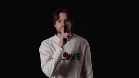 trendy young man shushing into the camera, shh gesture, wide, black background