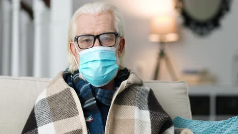 Primer-Plano-De-Un-Hombre-Enfermo-De-Pelo-Gris-Con-Una-Máscara-Médica-Y-En-El-Sofá-De-Casa-Y-Mirando-La-Cámara