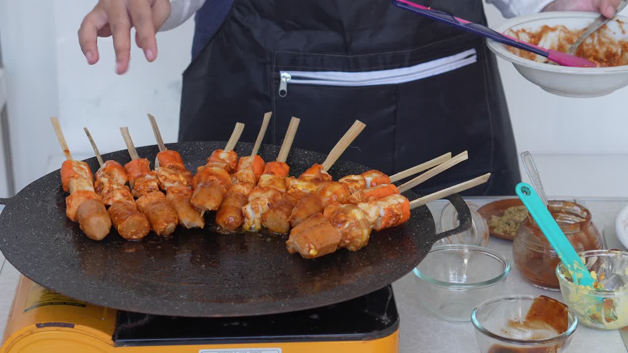 Nuori nainen esiliinassa kaatamassa maustetta ja levittämässä kastiketta kuumalla tulella grillattujen Suki Satay -vartaiden päälle.