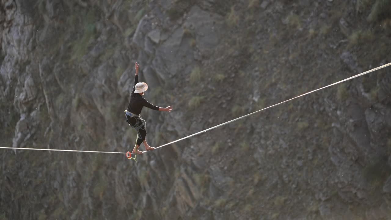 Slow-motion footage of an athlete balancing on a highline with safety harness, walking barefoot across a rope stretched in front of a rocky cliff