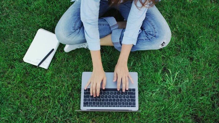 Topputsikt av kvinne som skriver på notebook-tastatur mens hun jobber på grønt gress i parken