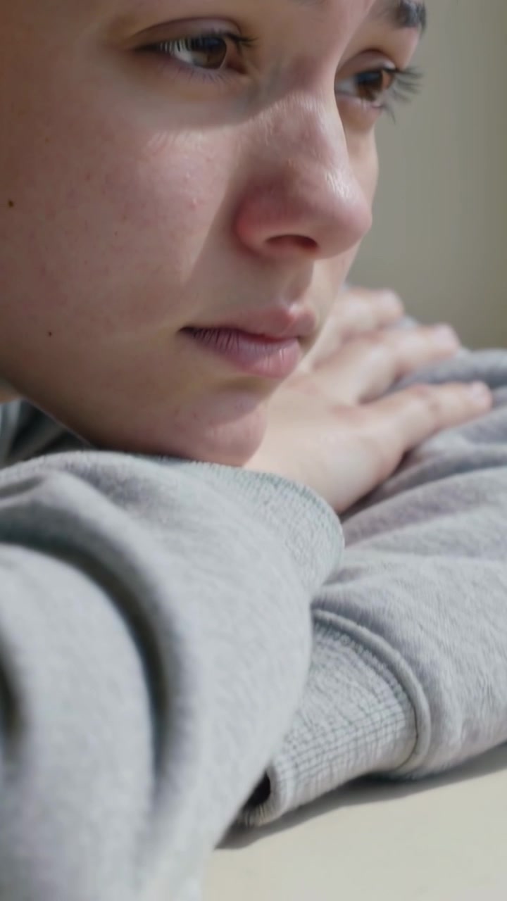Vertical video: Pulling back showing teen sad, chin resting on folded arms at table, grey top speck