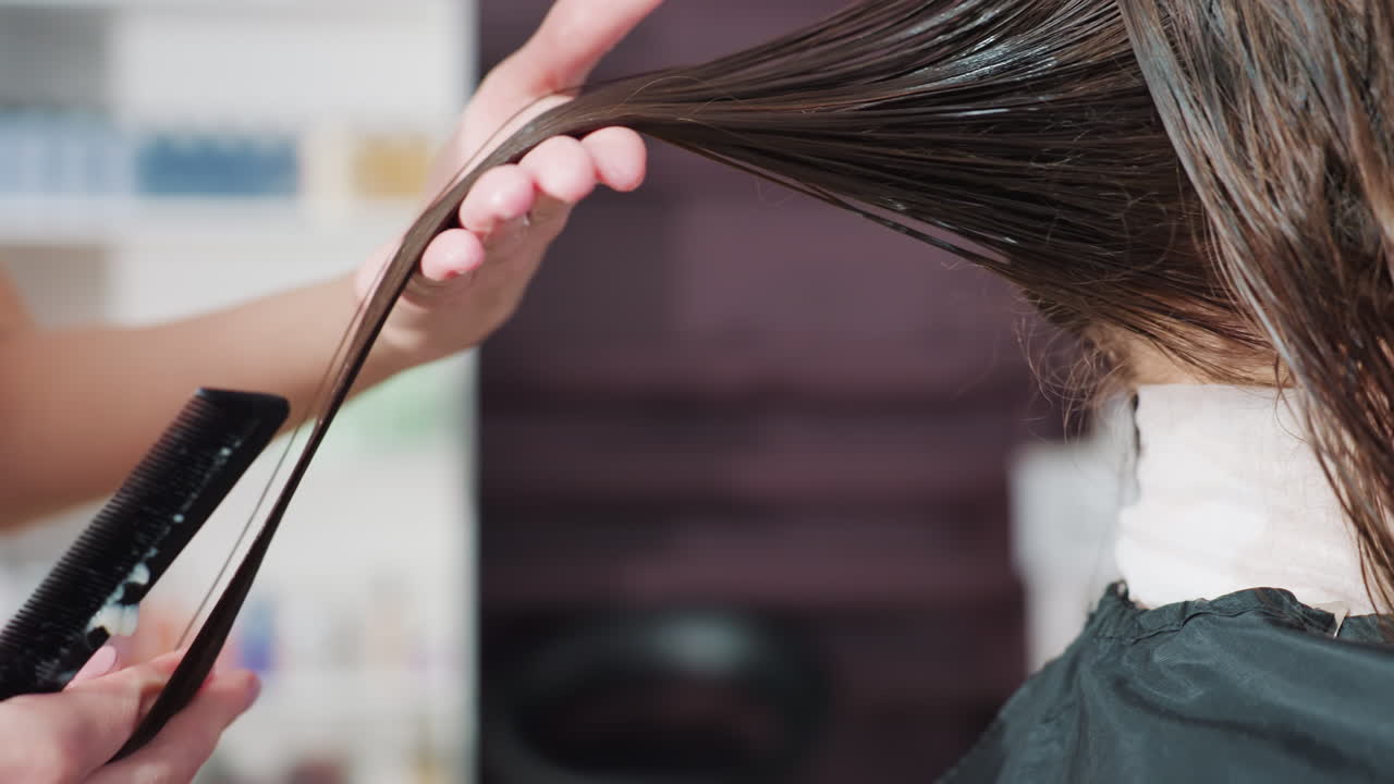 Lähikuva takaa, jossa stylisti muotoilee pitkiä, märkiä hiuksia saksilla ja kammalla. Salonkihyllyt ovat sumeana taustalla. Tarkoituksellinen tekniikka luo tasapainoisen pituuden ja liikkeen, keskittynyt ammattilainen