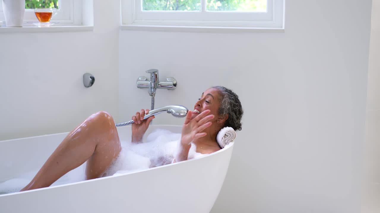 Senior African woman settling on towel in bathtub holding shower head, waving and smiling, relaxing