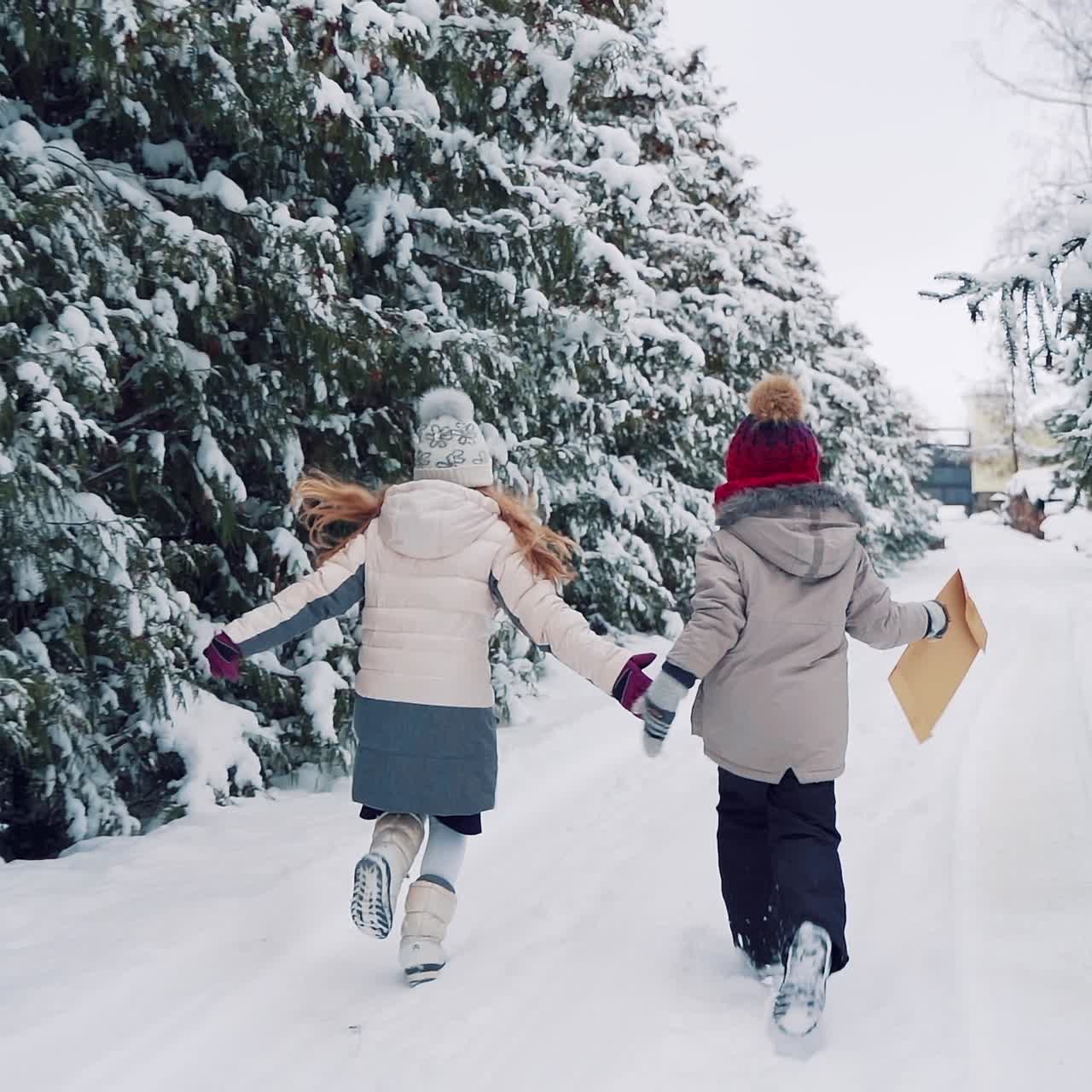 Children with their backs are running along the snowy road and holding each other's hands on the background of coniferous forest on two sides. The little boy is holding a brown envelope in his hand