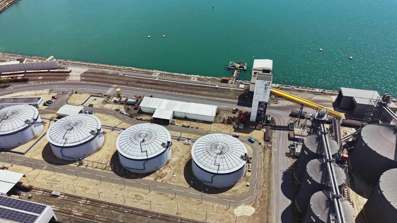 Large storage tanks and industrial facilities next to water body in a coastal area during daytime with clear blue skies above